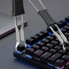 Primer plano de una mano usando un extractor de keycaps plateado para quitar una tecla de un teclado mecánico negro.