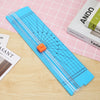 A blue portable manual paper trimmer with a clear ruler and orange guide on a wooden surface.