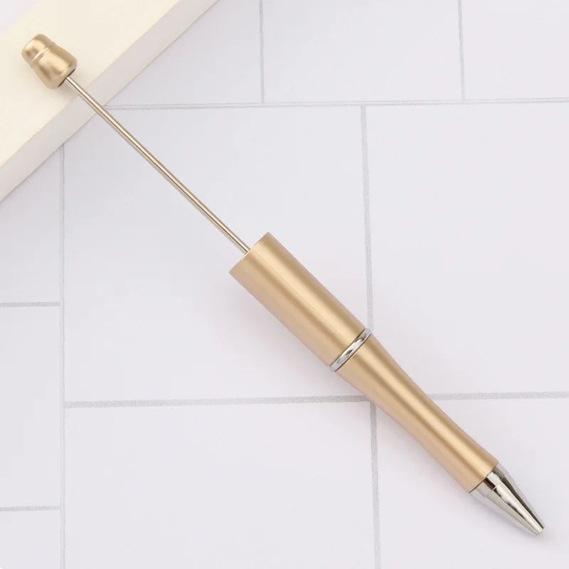 A gold-colored DIY beadable pen is shown lying on a white tiled surface.