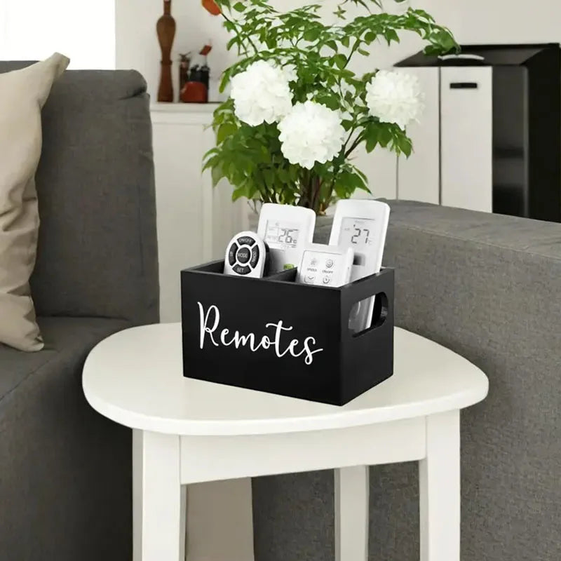 A black storage box with dividers holding various remote controls and small electronic devices on a white table.
