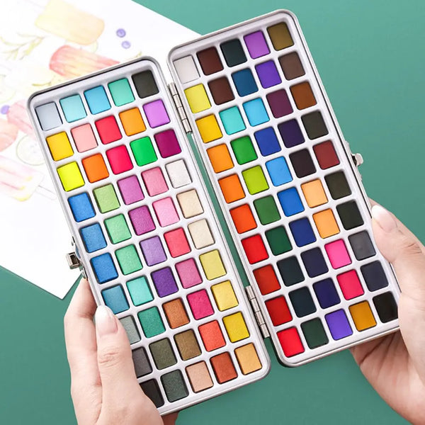 A hand holds an open palette of solid watercolor paints featuring various colors arranged in a grid.