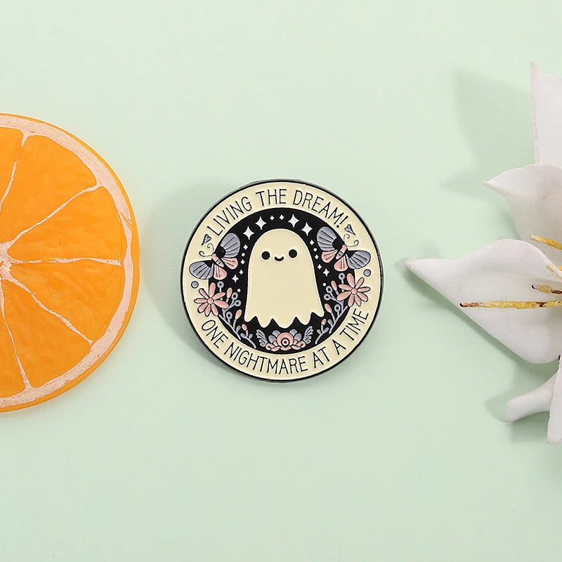 A round enamel pin featuring a cute ghost design with bats and flowers, surrounded by text that reads Living the Dream at a T