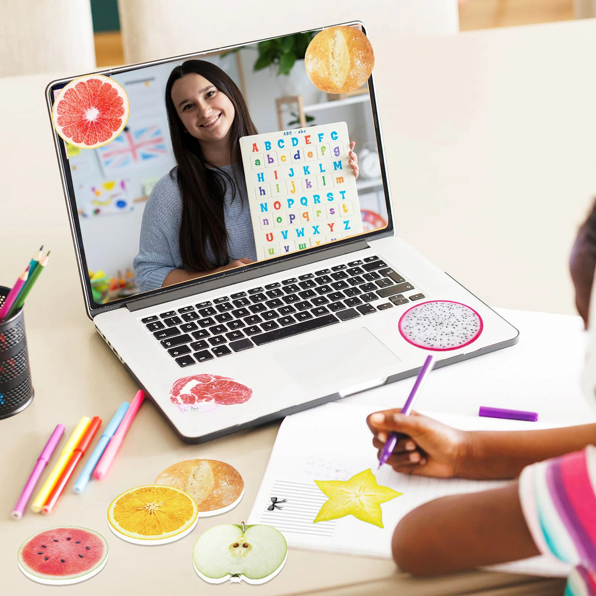 A laptop displaying an alphabet grid is surrounded by various fruit-shaped sticky notes on a desk.