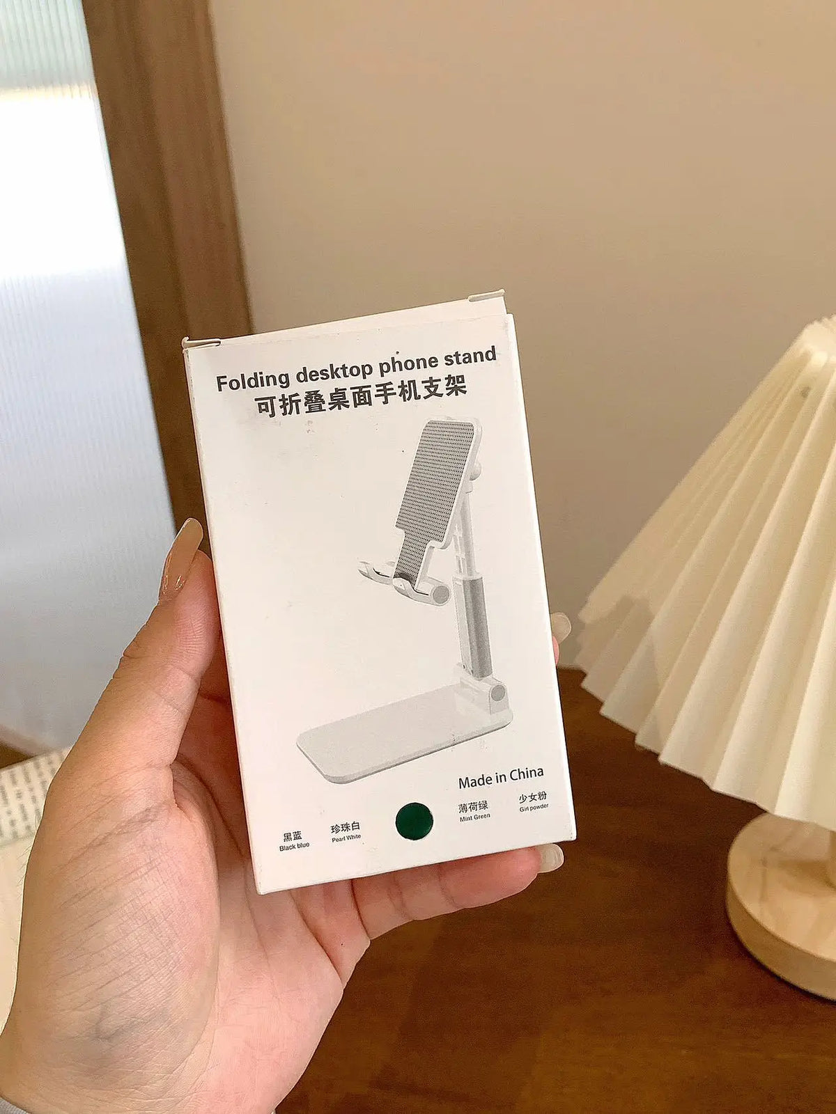A white folding desktop phone stand is shown in a hand, demonstrating its adjustable design.