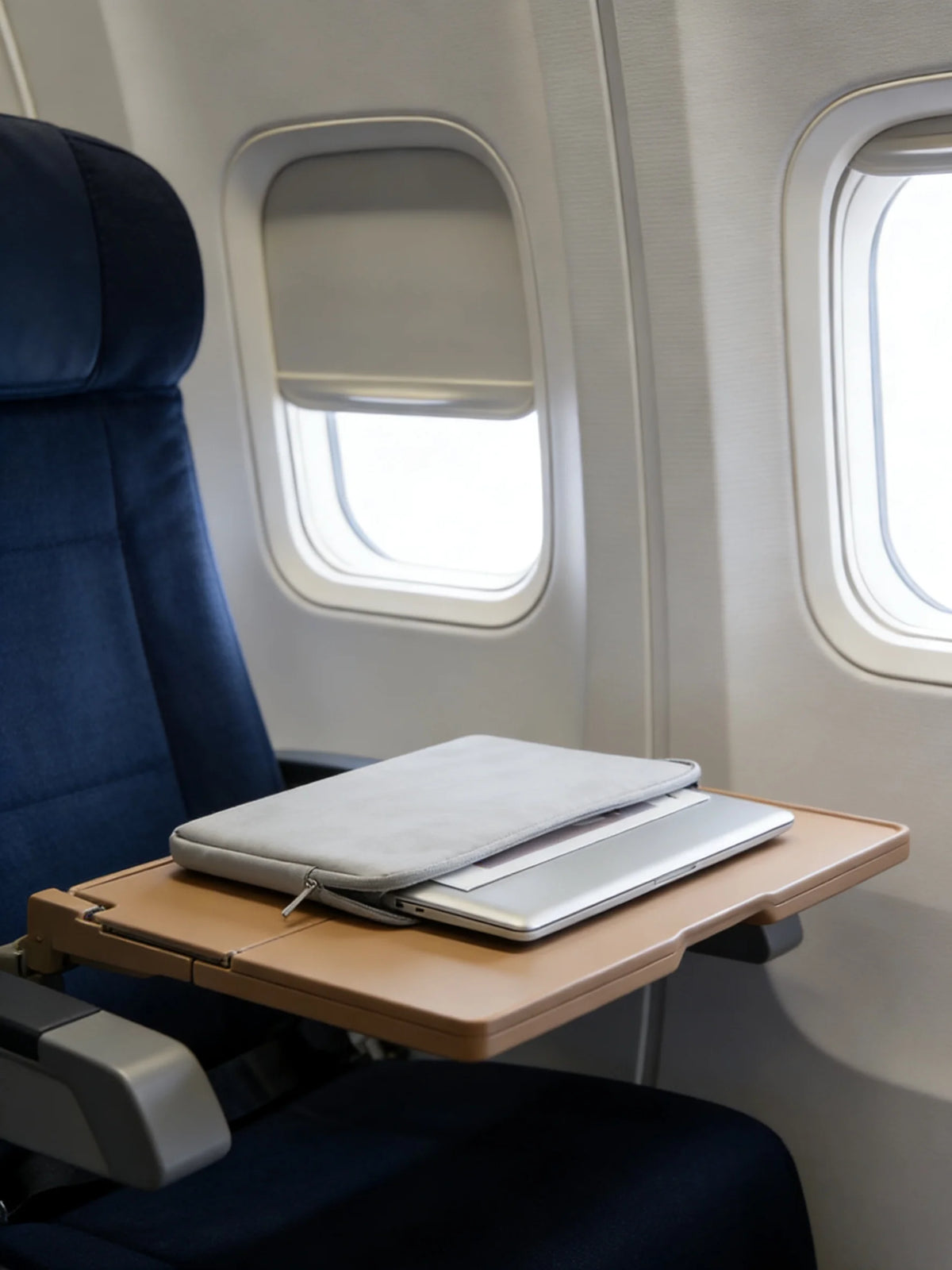 A laptop sleeve is placed on a tray table in an airplane seat, next to a window.