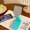 A turquoise adjustable foldable phone and tablet stand resting on a white notebook next to a wooden table.