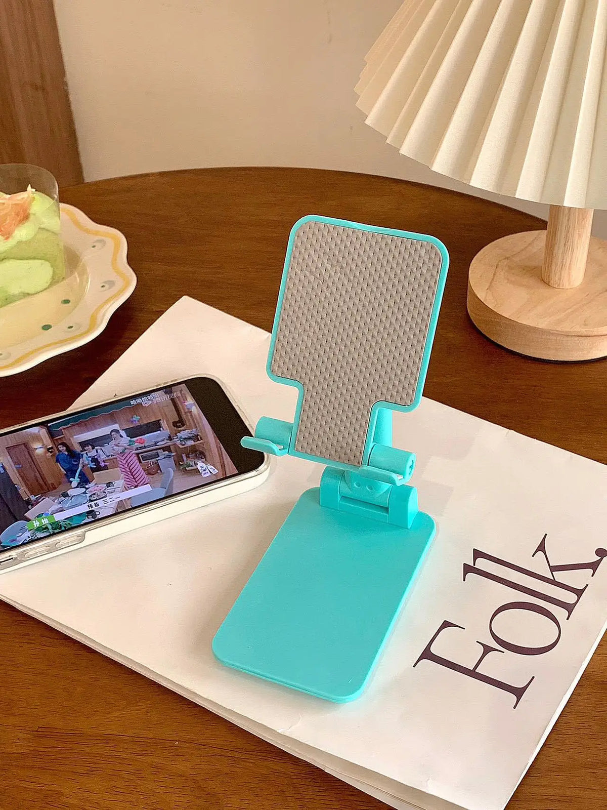 A turquoise adjustable foldable phone and tablet stand resting on a white notebook next to a wooden table.