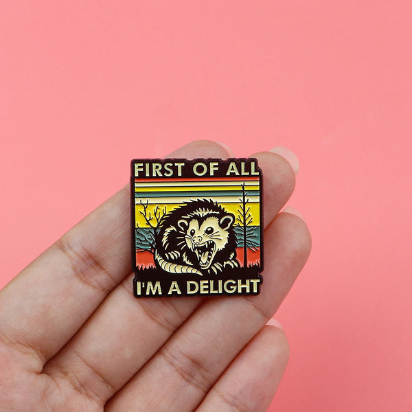 An enamel pin featuring a stylized, fierce-looking animal face with text that reads First of All I'm a delight.