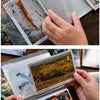 A hand holds an A6 photo storage booklet displaying a landscape painting.