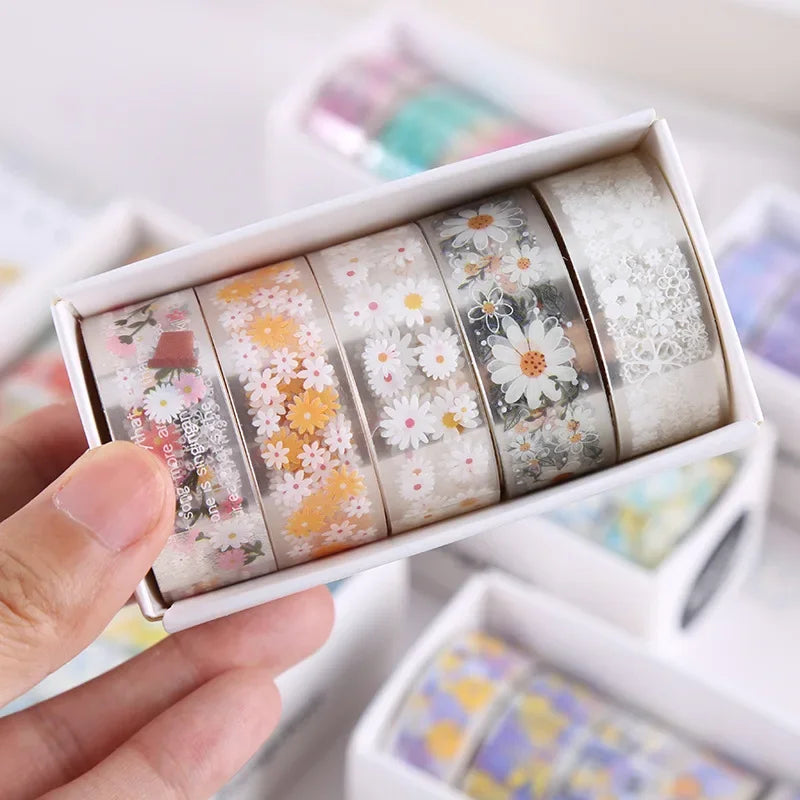 A hand holds a box of decorative transparent washi tape rolls with floral patterns.