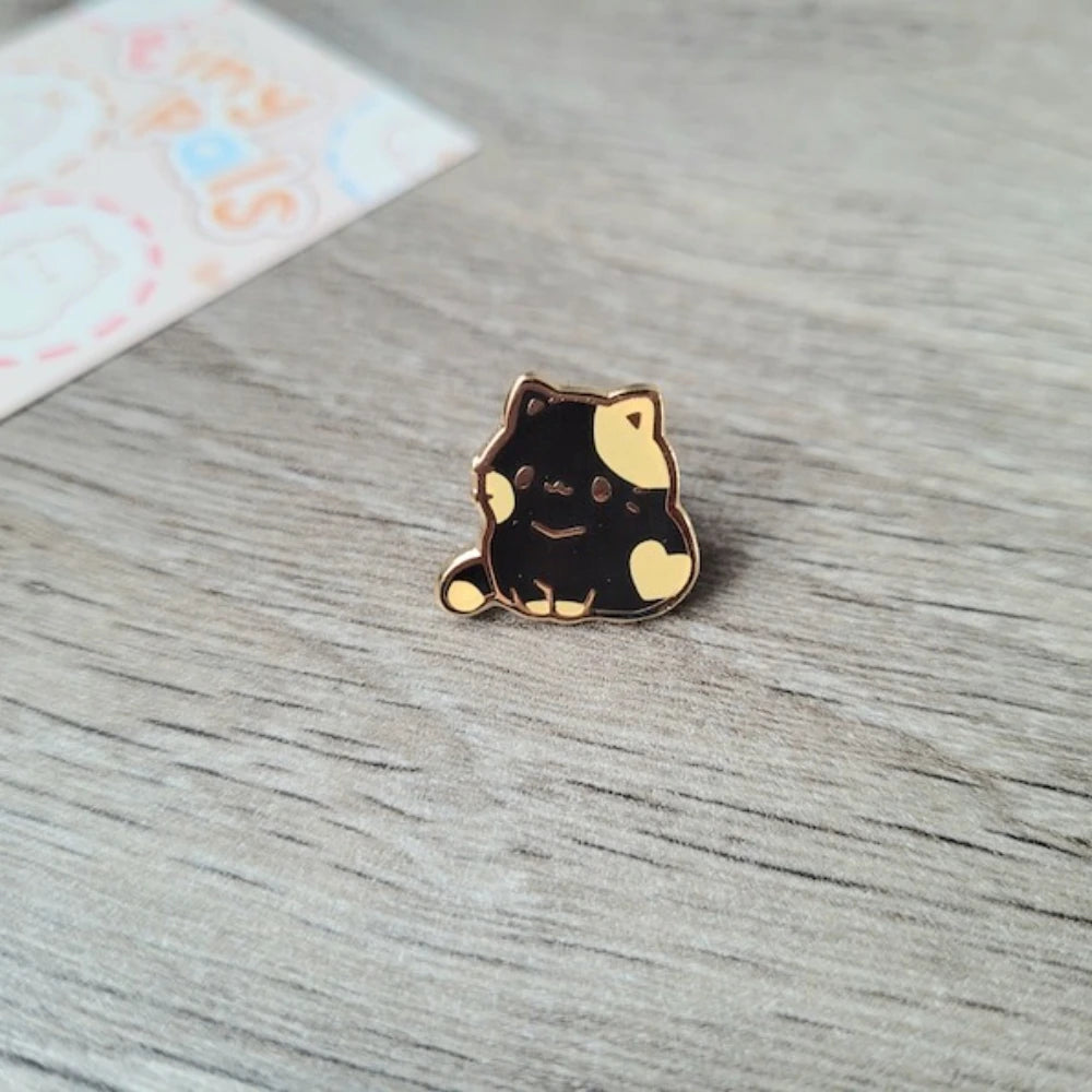 Un pequeño pin de esmalte de color dorado con forma de un gato negro y amarillo adorable, descansando sobre una superficie de