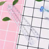 A clear acrylic folding ruler set with markings and a small blue gemstone is displayed on a pink grid background.