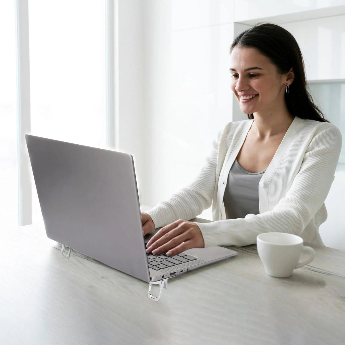 Une femme travaille sur un ordinateur portable avec un cardigan blanc et une tasse blanche sur un bureau en bois clair.