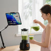 A woman prepares a green smoothie using an adjustable tablet stand and blender on a wooden table.