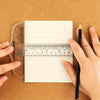 Hands hold a pencil and an acrylic T-square ruler set on brown paper for measuring or crafting.