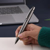 A hand holds a silver metal ballpoint pen against a white surface with a laptop in the background.