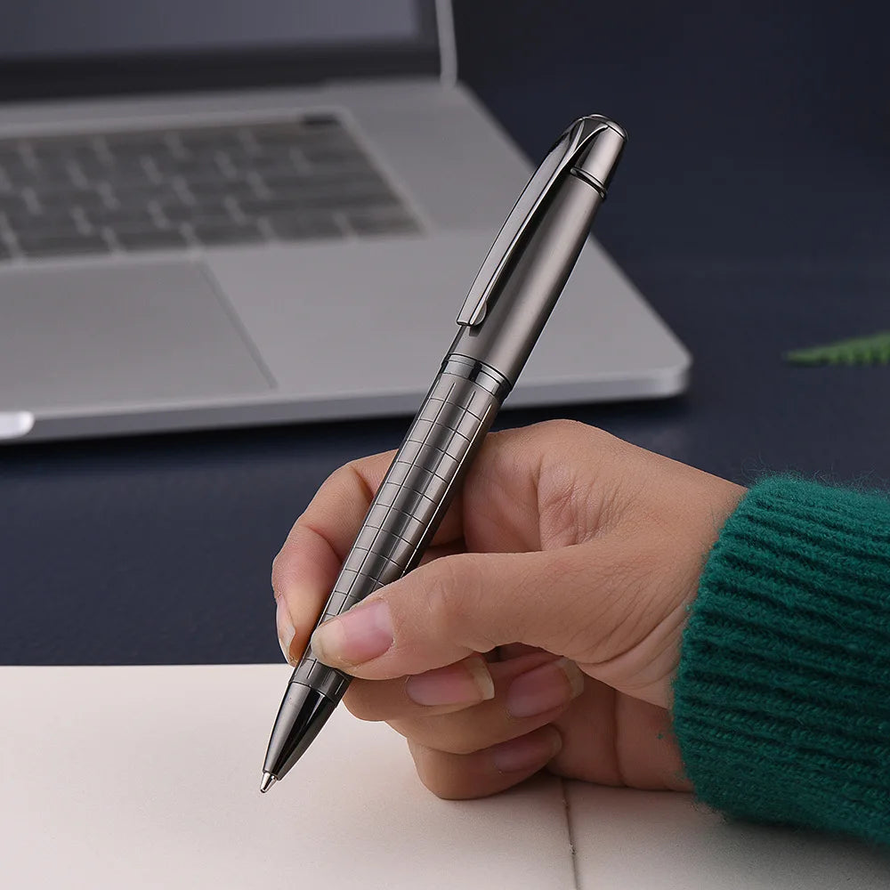 A hand holds a silver metal ballpoint pen against a white surface with a laptop in the background.