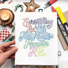 A hand holds a sheet of paper with colorful script writing on it, surrounded by art supplies.