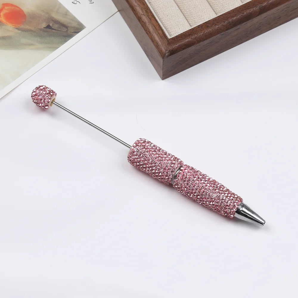 A silver pen accessory with a pink rhinestone tip and decorative beadwork is shown on a white background.