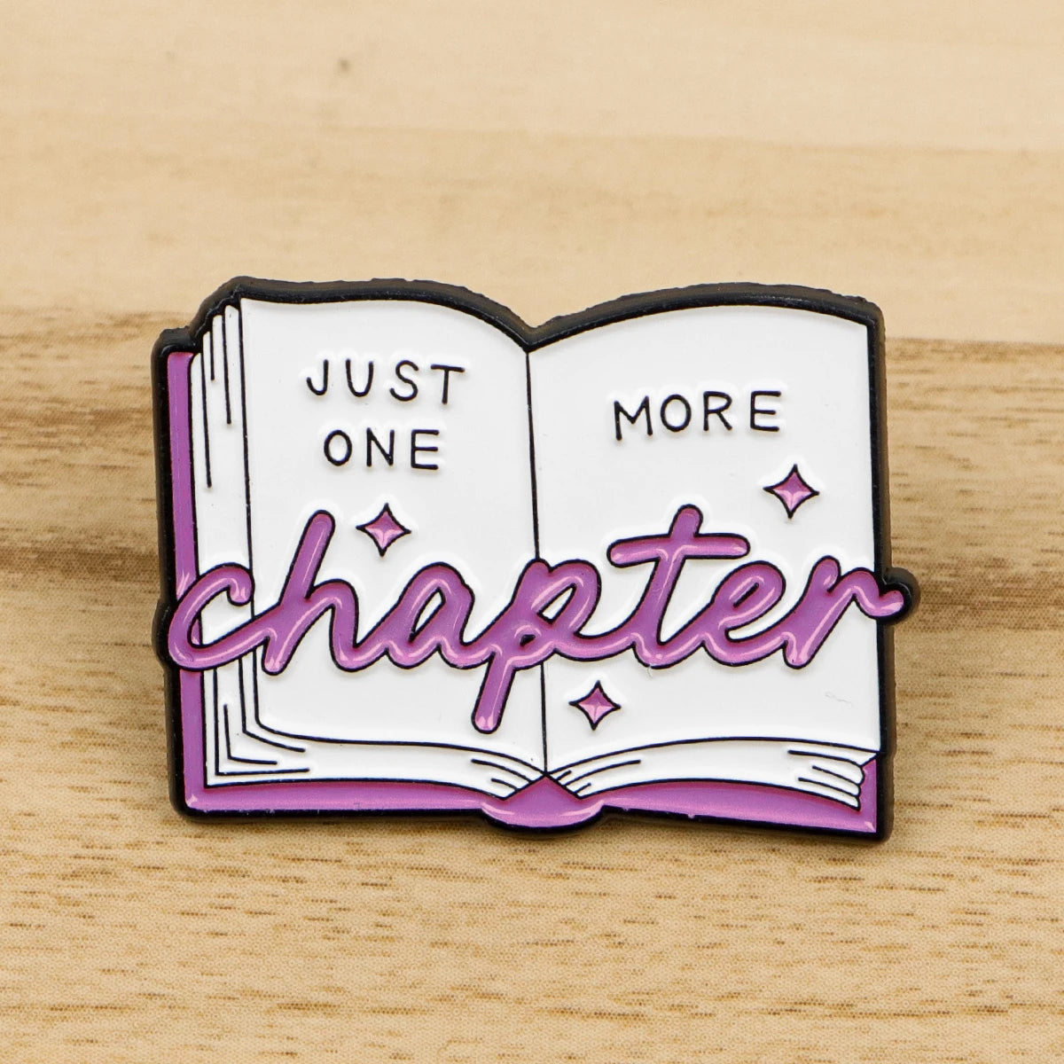 A lapel pin featuring an open book design with the text Just One More Chapter in purple and white enamel.