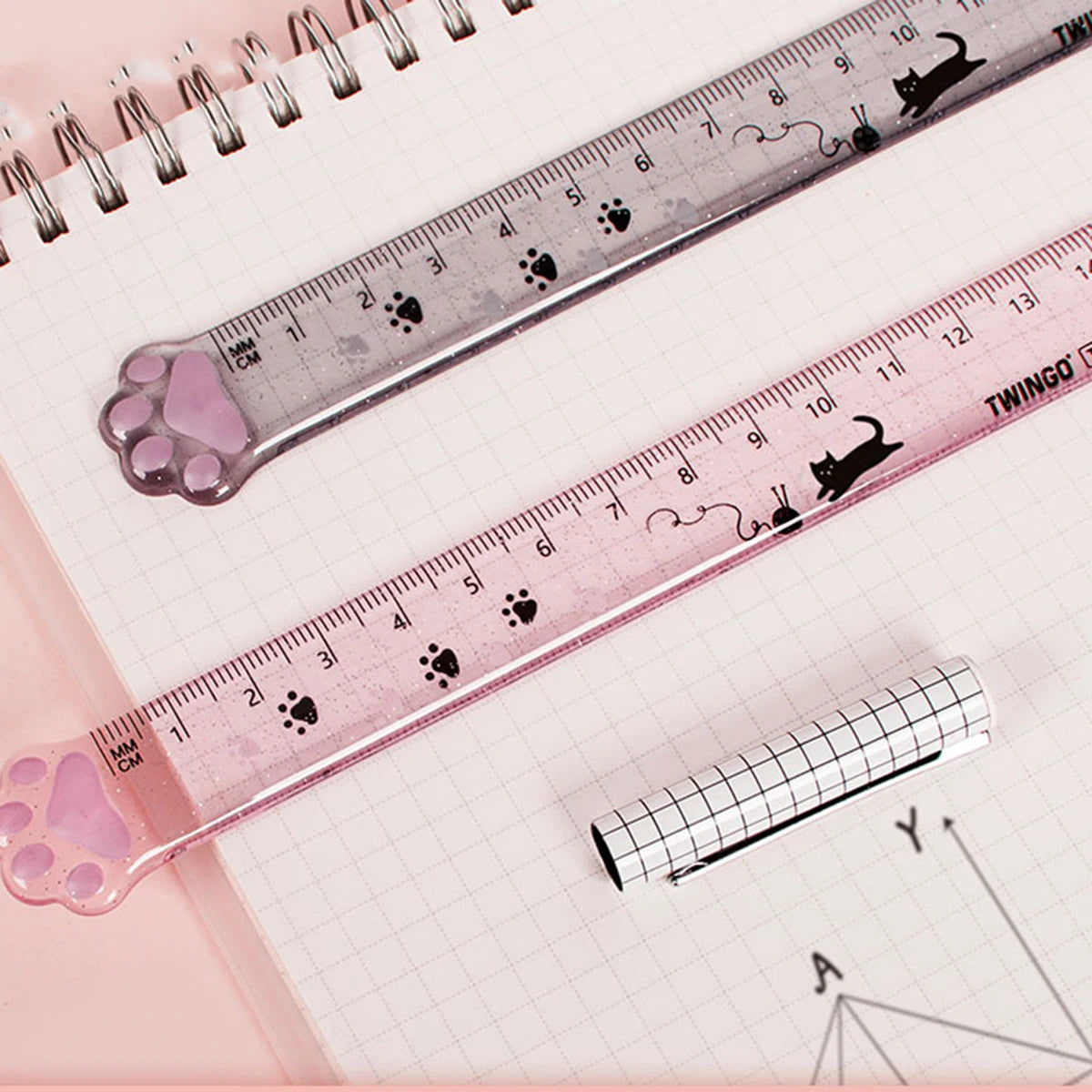 A pink plastic measuring tool with cat paw markings and a ruler scale is shown on lined paper.