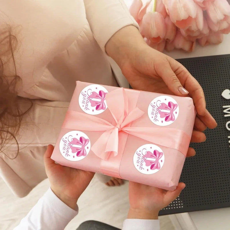 Eine Hand hält eine rosa Danksagungsstickerbox zum Scrapbooking mit rosa Schleife vor dunklem Hintergrund.