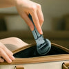 A hand is using a 3-in-1 magnetic phone holder and selfie stick attached to a tan leather handbag.