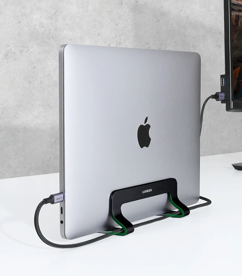 A silver laptop is supported by a black vertical stand with green cables on a white surface.