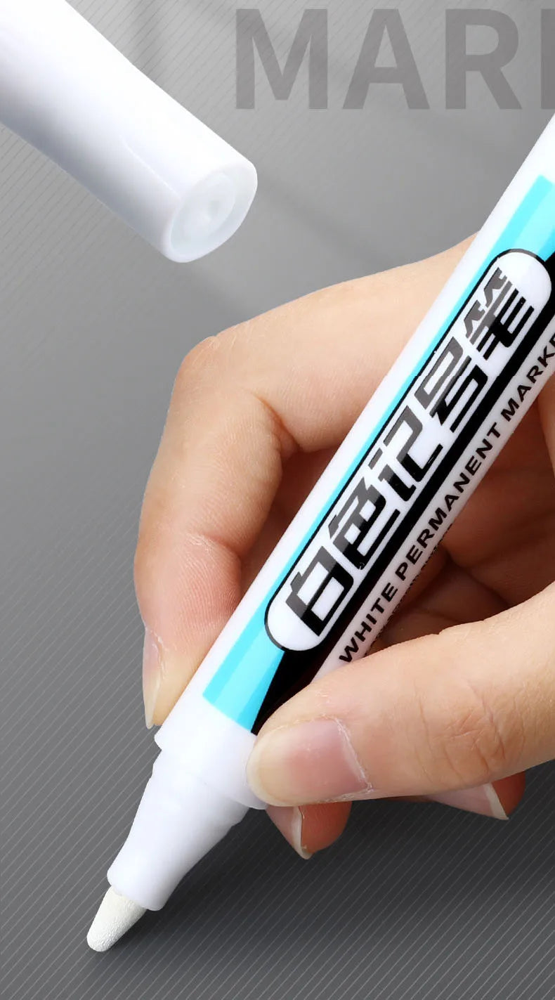A hand holds a white permanent paint marker pen with blue accents, showing the tip and text.