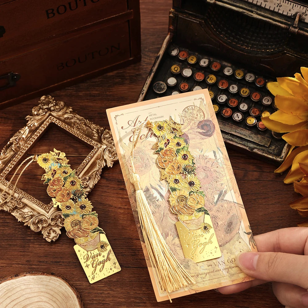 A hand holds a vintage artistic metal bookmark with a gold tassel resting on a wooden surface.