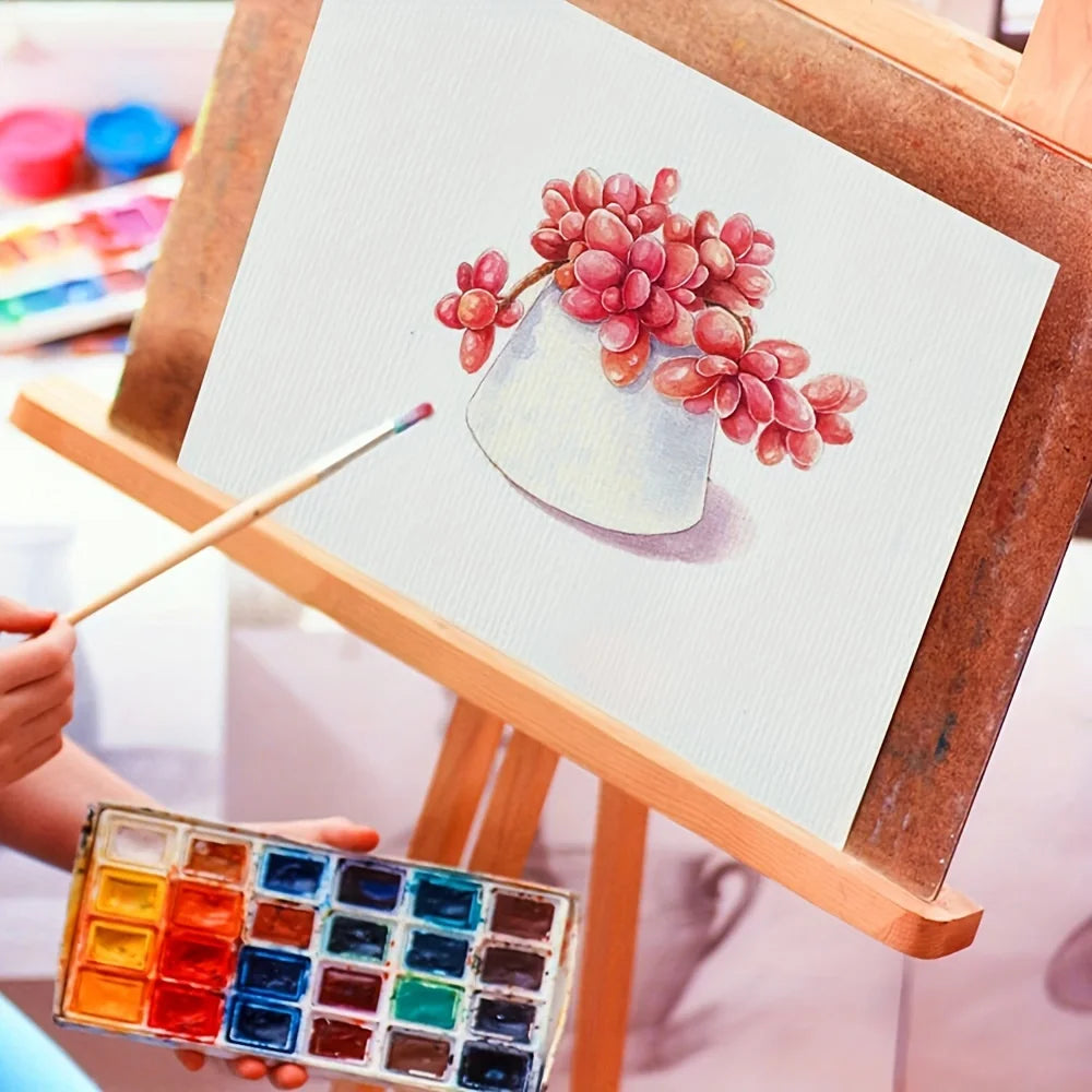 A hand paints a watercolor painting of pink and red grapes on a canvas using a paintbrush and a palette.