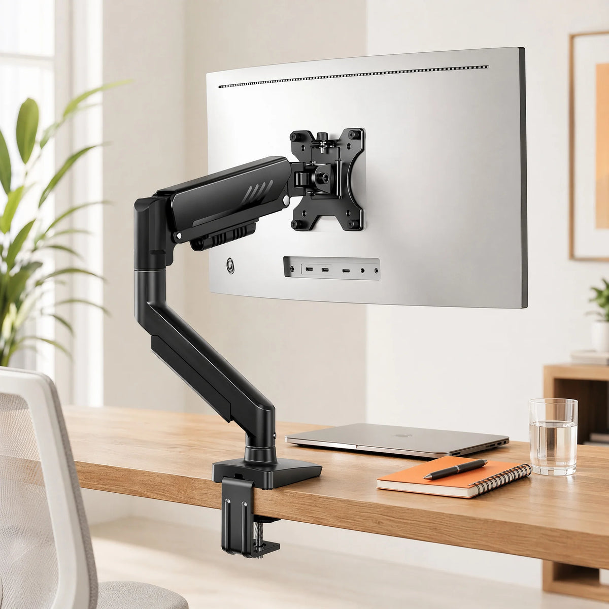 A black monitor arm is mounted on a wooden desk, positioned above a laptop and notebook.