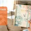 A spiral bound planner with monthly tabs showing a floral watercolor design and dimensions.