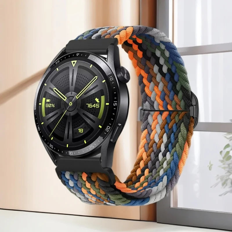 A black smartwatch with a woven orange and gray nylon strap is displayed next to a window.