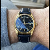 A close-up photo of a men's watch with a gold-toned case and black leather strap on a wrist.