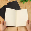 A blank A6 sketchbook with cream-colored pages is open, resting on a wooden surface.