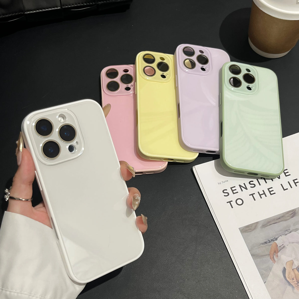 A hand holds a white iPhone case next to several colored, clear cases arranged on a dark surface.