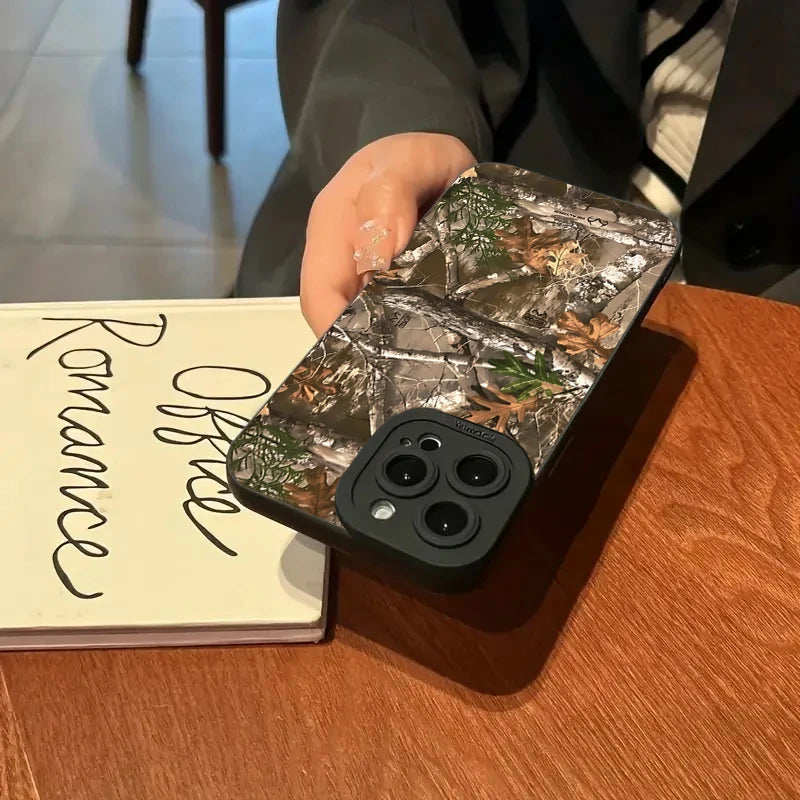Une main tient une coque de téléphone en silicone camouflage forêt pour iPhone posée sur une table en bois à côté d'un carnet