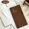 A brown slim weekly planner journal with a gold logo is shown next to loose pages of a white planner on a white fabric backgr