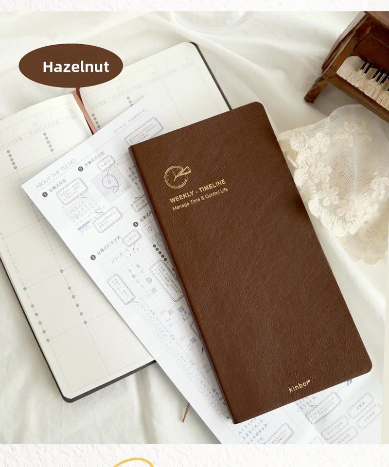 A brown slim weekly planner journal with a gold logo is shown next to loose pages of a white planner on a white fabric backgr