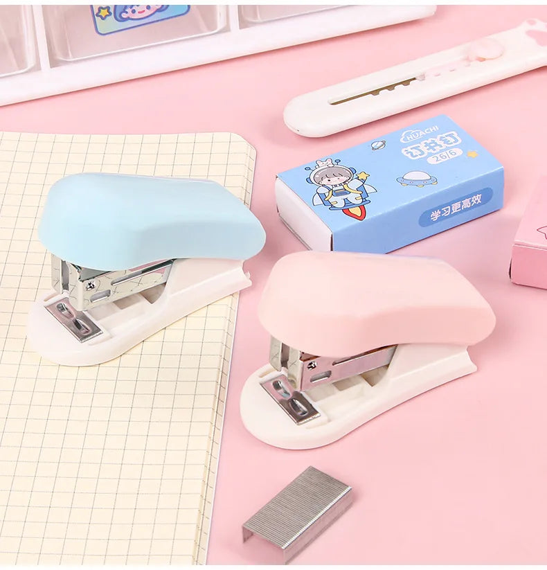 A set of cartoon mini staplers in light blue and pink, displayed on a pink background with stationery.