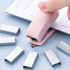A pink compact mini stapler kit with metal staples is shown on a light blue surface.