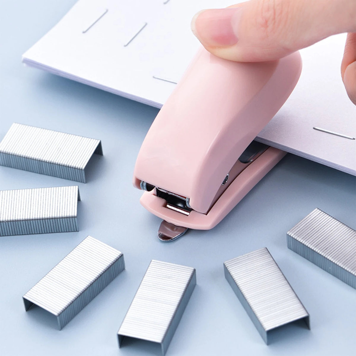 A pink compact mini stapler kit with metal staples is shown on a light blue surface.