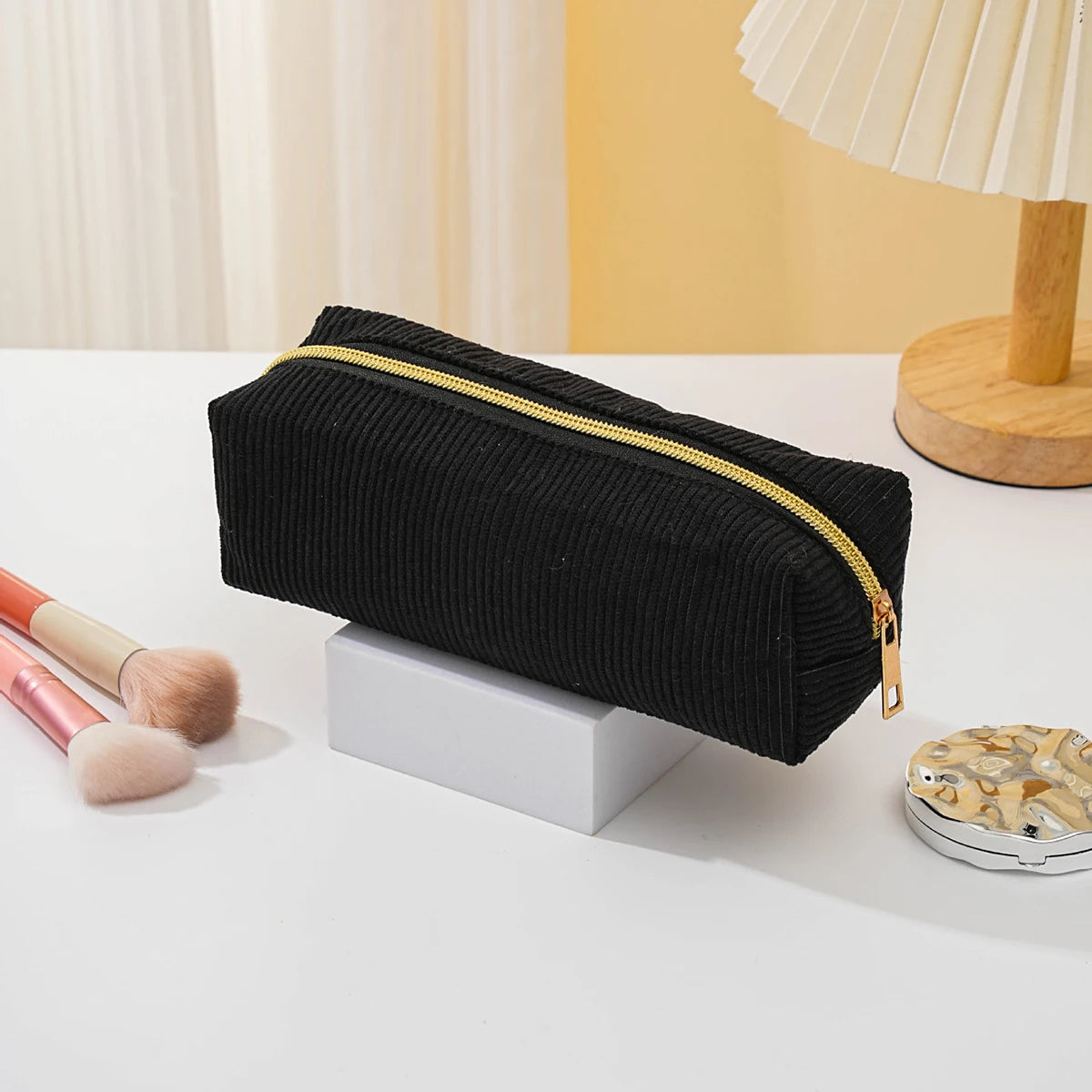 Black zippered pencil case resting on a white surface next to a makeup brush and a wooden lamp base.