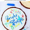 A round cake decorated with bright blue and yellow lettering, surrounded by an acrylic marker pen.