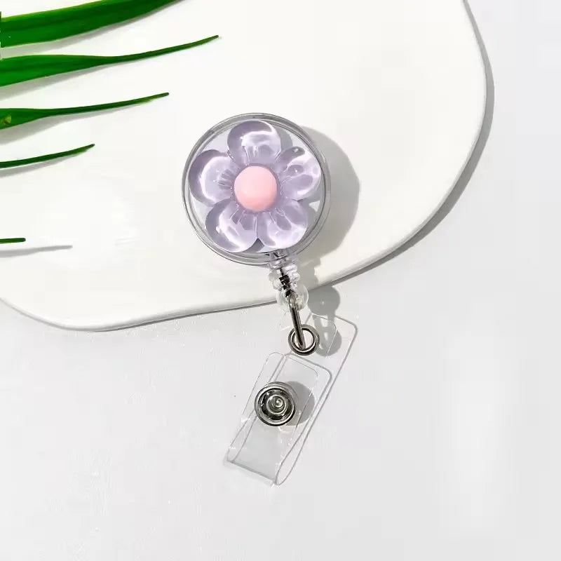 A clear plastic retractable ID badge holder with a purple floral pull buckle and a pink charm