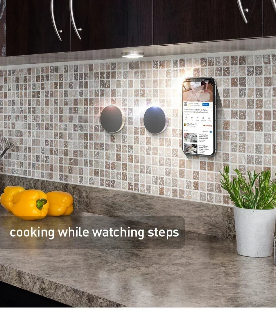 A magnetic phone mount is attached to a kitchen backsplash, showing a smartphone displaying a video.