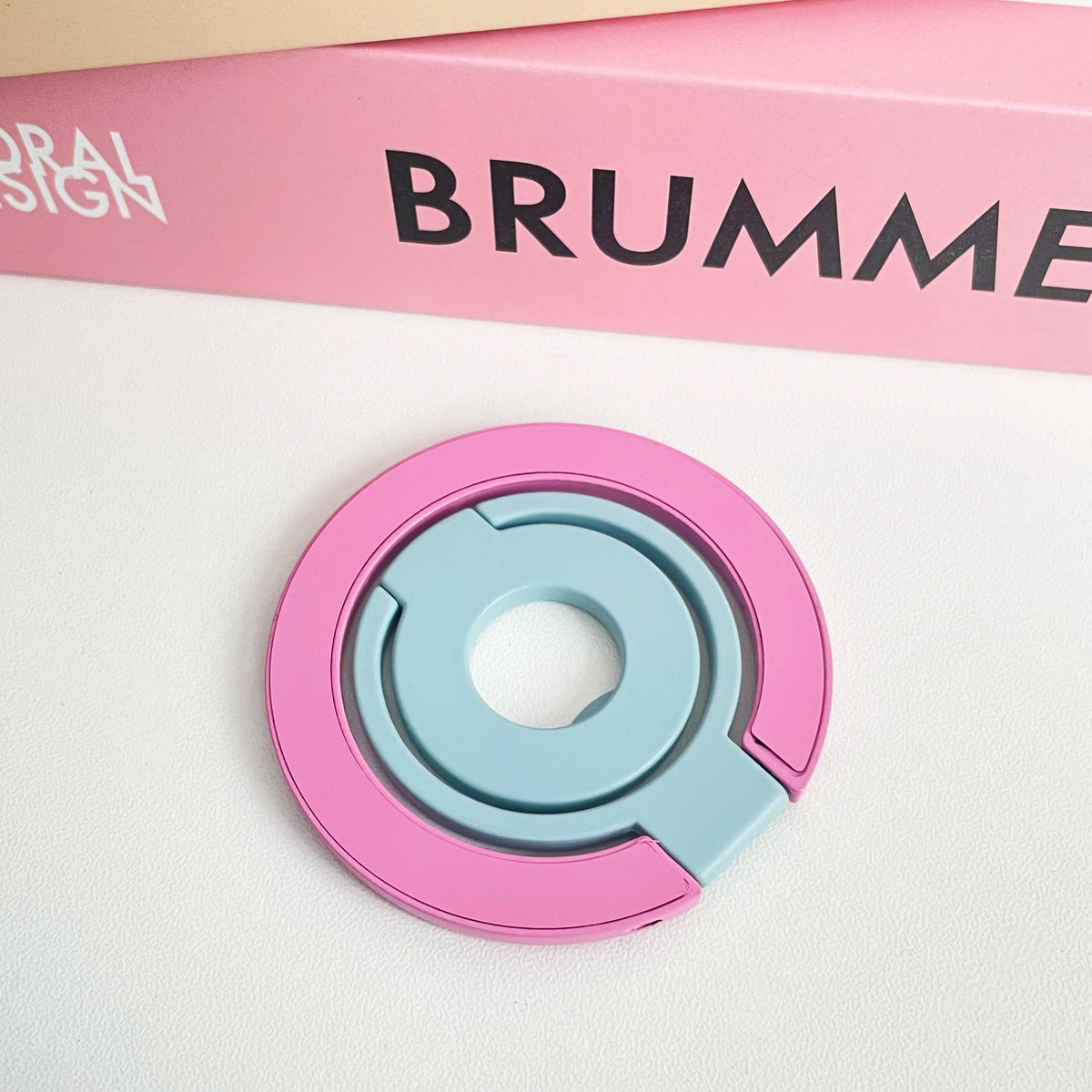 A pink and blue magnetic phone grip and stand with a foldable ring resting on a white surface.