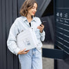 Eine Frau in einem Jeanshemd hält ein weißes Polybeutelpaket und schaut auf ihr Smartphone.