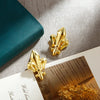 A pair of gold-colored, iris-shaped metal binder clips resting on a light surface next to a teal object.
